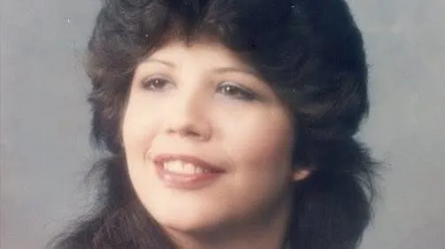 Angie Buck smiling, with feathered dark brown hair, in a portrait against a gray background